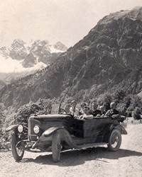 Mehrere Personen sitzen in einem historischen Oldtimer auf einer Straße im Brandnertal. Im Hintergrund ist eine imposante Berglandschaft zu sehen.