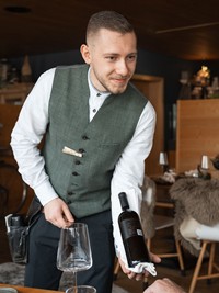 Ein Mitarbeiter des Hotels Valavier serviert einem Gast eine Flasche Wein in einem gemütlichen Restaurant mit rustikalem Ambiente.