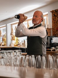 Ein Mitarbeiter des Hotels Valavier mixt hinter der Bar einen Drink, während zahlreiche Gläser und edle Spirituosen die gemütliche Hotelbar schmücken.