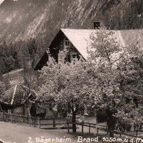 Eine historische Aufnahme zeigt das Jägerheim in Brand vor der beeindruckenden Bergkulisse des Brandnertals und umgeben von Bäumen.