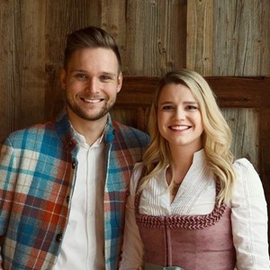 Fabian und Santina präsentieren sich lächelnd als Gastgeber des Hotels Valavier. Im Hintergrund sorgt eine rustikale Holzwand für eine warme und gemütliche Atmosphäre.