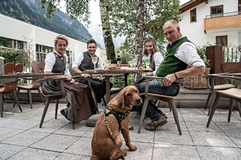 Gastgeberfamilie des Hotels Valavier sitzt gemeinsam mit ihrem Hund auf der Terrasse und genießt eine gesellige Runde inmitten der Berglandschaft.