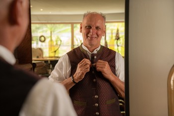 Gastgeber Edi vom Hotel Valavier richtet vor einem Spiegel seine traditionelle Trachtenweste und lächelt dabei.