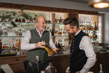 Der Barkeeper hält eine Flasche und tauscht sich mit dem Gastgeber des Hotels Valavier aus, der ihm aufmerksam zuhört. Im Hintergrund sind die stilvolle Hotelbar mit Regalen voller Spirituosen, natürlichen Holzelementen und großen Fenstern mit Blick auf die umliegende Berglandschaft zu sehen.