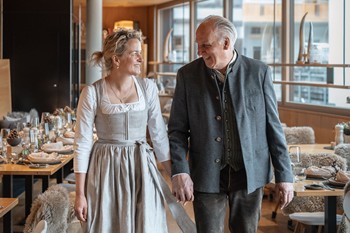 Die Gastgeber Kathi und Edi gehen Hand in Hand durch den Restaurantbereich des Hotels Valavier und lächeln sich dabei an. Im Hintergrund sind gedeckte Tische, gemütliche Sitzplätze und große Fenster mit Blick auf die Umgebung zu sehen.