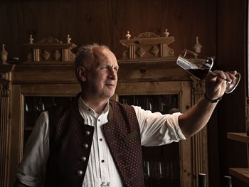 Edi vom Hotel Valavier präsentiert ein Glas Rotwein in gemütlicher Umgebung. Der traditionelle Kamin im Hintergrund verleiht dem Raum eine warme und einladende Atmosphäre.