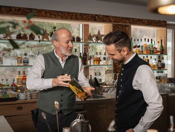 Der Barkeeper hält eine Flasche und tauscht sich mit dem Gastgeber des Hotels Valavier aus, der ihm aufmerksam zuhört. Im Hintergrund sind die stilvolle Hotelbar mit Regalen voller Spirituosen, natürlichen Holzelementen und großen Fenstern mit Blick auf die umliegende Berglandschaft zu sehen.