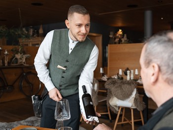 Gastfreundschaft und Service im Hotel Valavier Ein Mitarbeiter des Hotels Valavier serviert einem Gast eine Flasche Wein in einem gemütlichen Restaurant mit rustikalem Ambiente.