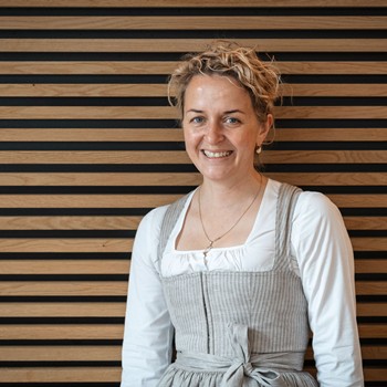 Kathi, Gastgeberin des Hotels Valavier, lächelt freundlich in die Kamera. Sie trägt eine traditionelle Tracht und steht vor einer warm gestalteten Holzwand.