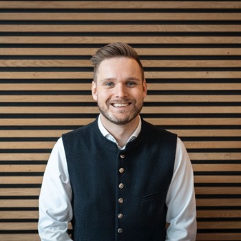 Fabian, Gastgeber im Hotel Valavier, lächelt freundlich in die Kamera. Er trägt eine traditionelle Weste und steht vor einer Wand aus warmen Holzelementen.