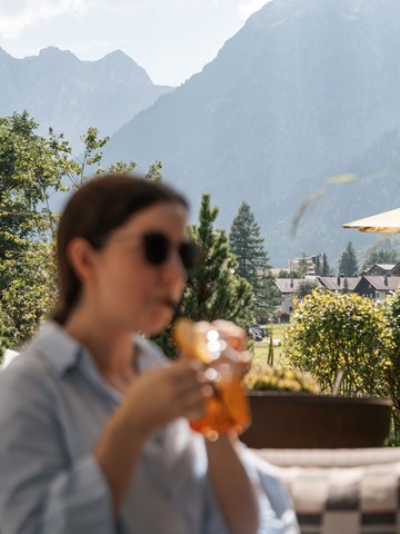 Zeit für Erfrischung Eine Frau genießt ein erfrischendes Getränk auf der Terrasse. Im Hintergrund erstrecken sich die Berge und der Ort Brand im Brandnertal.