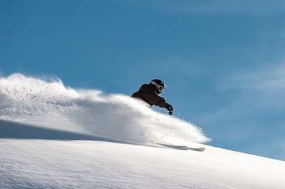 Ein Skifahrer fährt durch tiefen Pulverschnee und wirbelt dabei eine große Schneewolke auf. Der klare blaue Himmel unterstreicht die winterliche Bergkulisse.