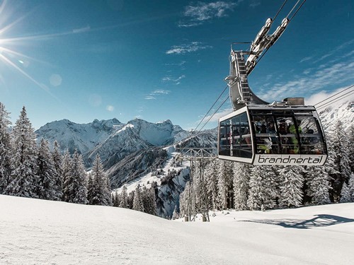 Eine Gondel der Bergbahnen Brandnertal schwebt über einer verschneiten Winterlandschaft. Dahinter erstreckt sich ein beeindruckendes Panorama aus schneebedeckten Bergen und tief verschneiten Wäldern.
