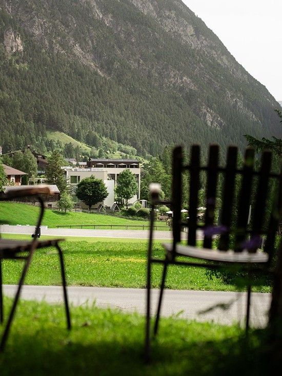 Zwei gemütliche Stühle stehen im grünen Garten und laden zum Entspannen ein. Dahinter erstrecken sich die Häuser von Brand und die imposante Berglandschaft des Brandnertals.