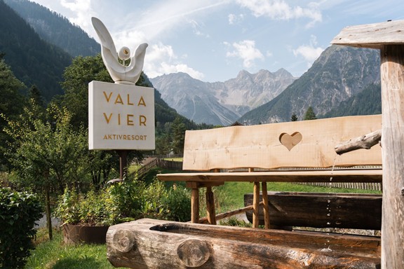 Das Valavier Hotelschild steht neben einer rustikalen Holzbank und einem traditionellen Holzbrunnen. Im Hintergrund eröffnet sich ein beeindruckendes Bergpanorama mit bewaldeten Hängen und markanten Gipfeln des Brandnertals.
