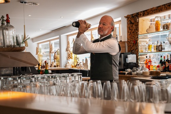 Ein Mitarbeiter des Hotels Valavier mixt hinter der Bar einen Drink, während zahlreiche Gläser und edle Spirituosen die gemütliche Hotelbar schmücken.
