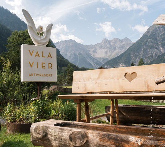 Bergpanorama zum Genießen Das Valavier Hotelschild steht neben einer rustikalen Holzbank und einem traditionellen Holzbrunnen. Im Hintergrund eröffnet sich ein beeindruckendes Bergpanorama mit bewaldeten Hängen und markanten Gipfeln des Brandnertals.