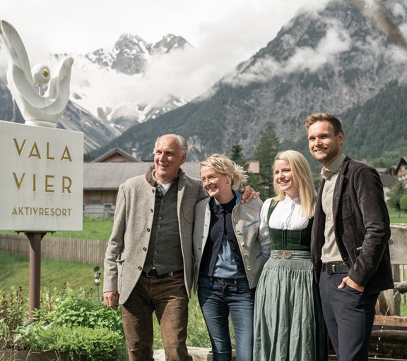 Die Gastgeberfamilie des Hotels Valavier steht neben dem Hotelschild und lächelt in die Ferne, während sich dahinter die schneebedeckten Berge des Brandnertals erheben.