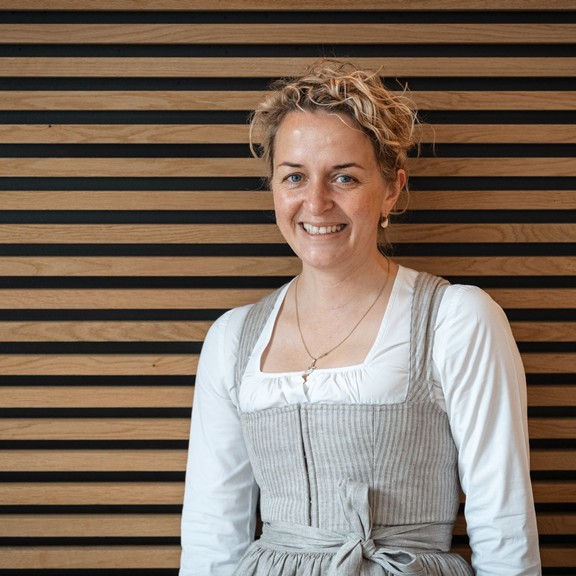 Kathi, Gastgeberin des Hotels Valavier, lächelt freundlich in die Kamera. Sie trägt eine traditionelle Tracht und steht vor einer warm gestalteten Holzwand.